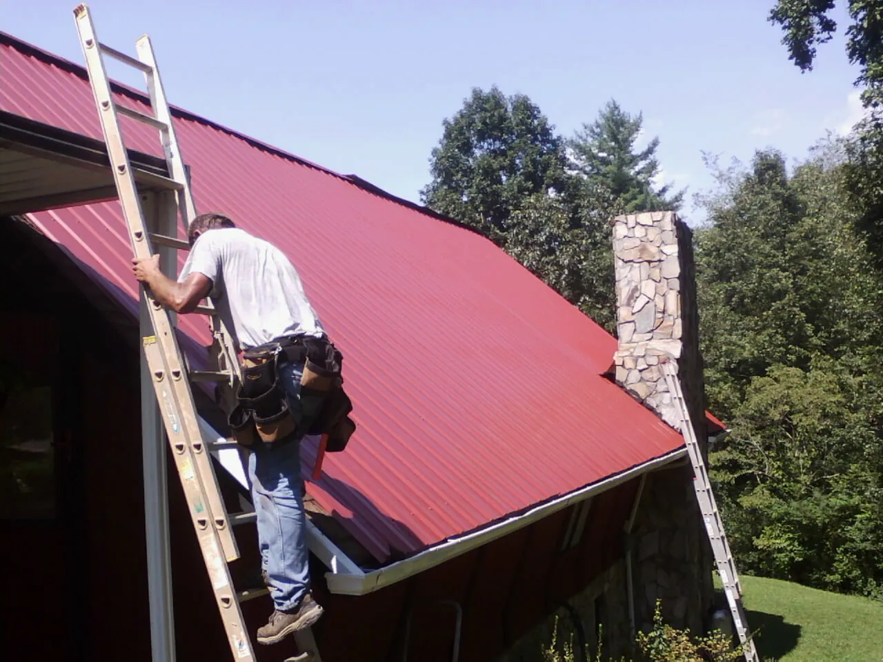 Roofer on a ladder beside a red metal roof and stone chimney — Soffit & Fascia Repair by Roofing Brothers in Walkersville